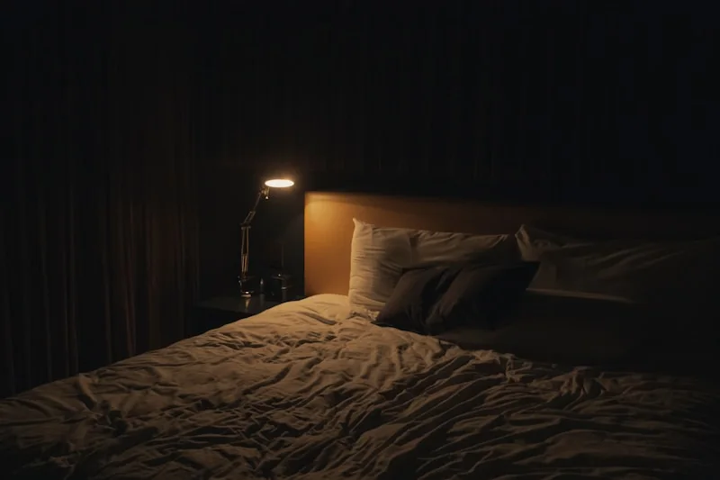 A dimly lit bedroom with a made bed and a small lamp glowing.